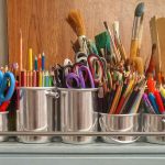 A diverse collection of art supplies in metallic buckets on a wooden table.