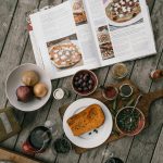 Flat lay showing a variety of cooking ingredients and an open cookbook on a rustic wooden table.