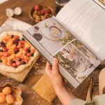 A person reads a cookbook while preparing a fruit tart on a wooden table.