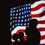 Silhouetted figures in front of a bright, wavy American flag create a patriotic scene.