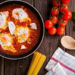 Delicious eggs in tomato sauce with basil, spaghetti, and fresh tomatoes on a wooden table.